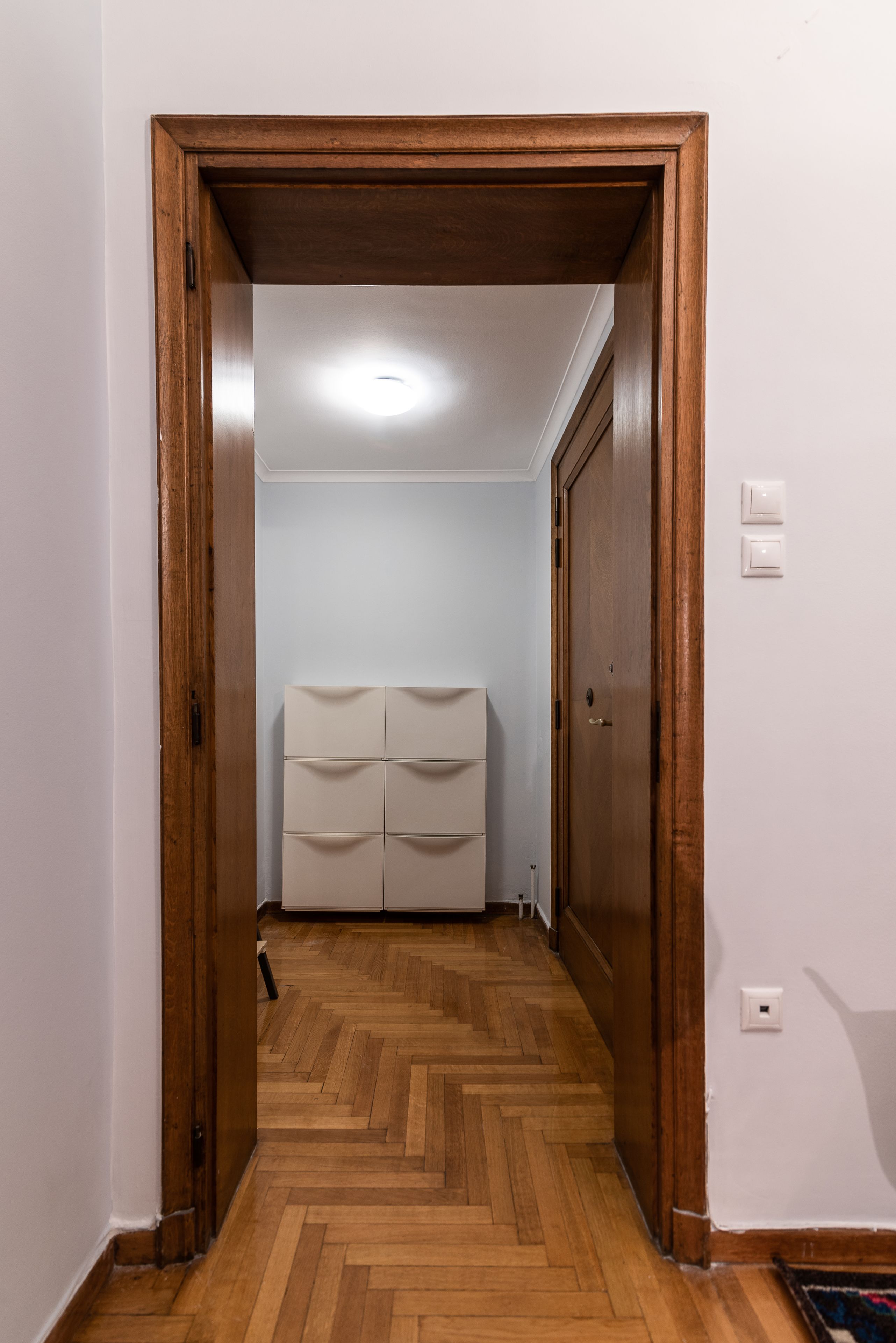 Egy folyosó, amelyben egy fa ajtókeret, egy fehér fal és halszálkamintás parketta található.