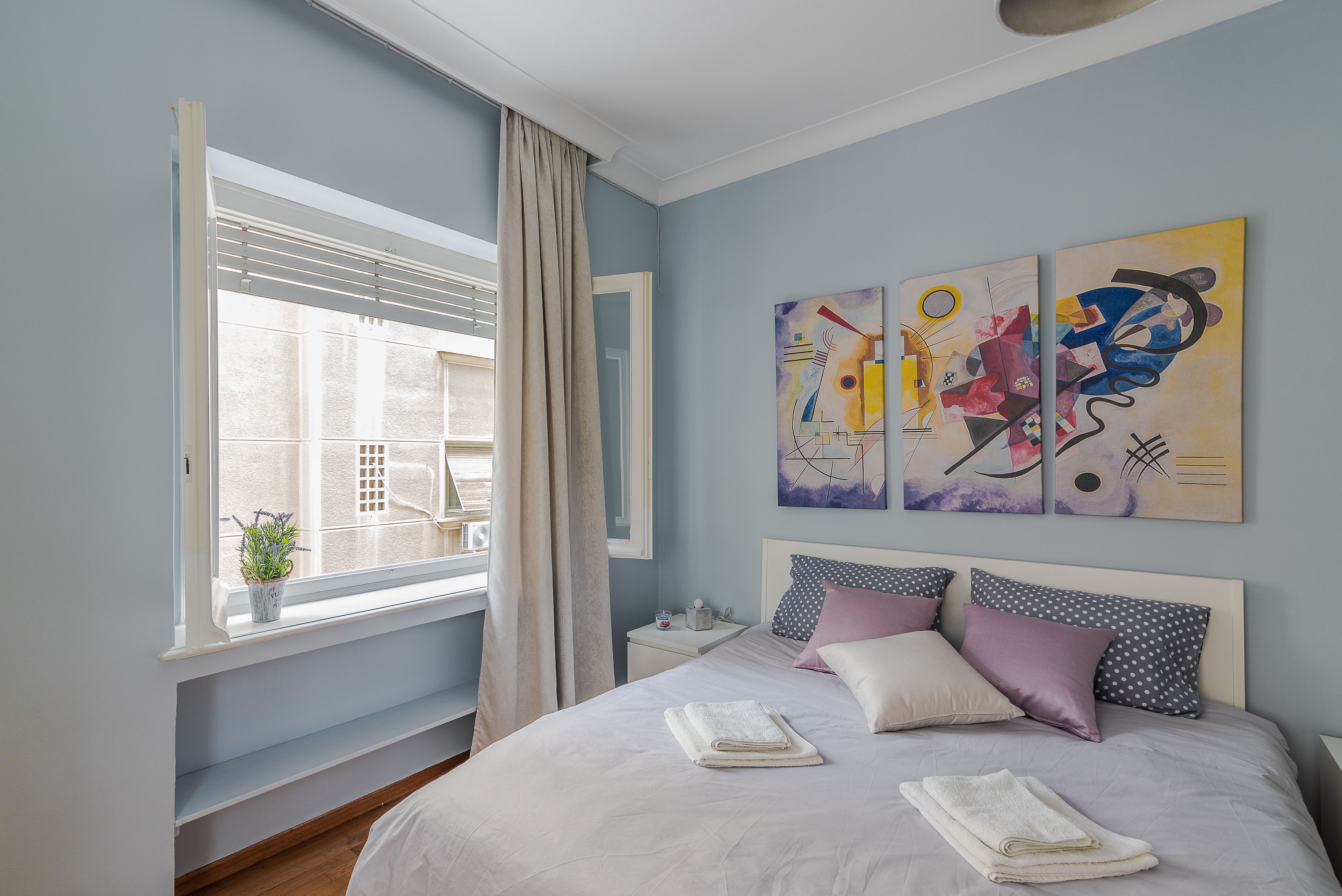 Een slaapkamer met een bed, een raam met gordijnen, een nachtkastje en abstracte wanddecoratie.