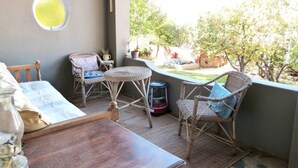 A balcony with wicker furniture, a round window, and a view of a backyard with trees and a fence. - Cosy 6 Person Cottage With Mountain View (Barrydale)