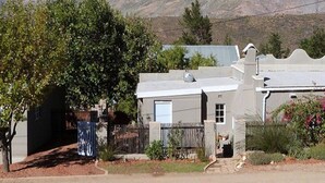 A residential house with a white exterior, a fenced yard, and a mountain backdrop. - Cosy 6 Person Cottage With Mountain View (Barrydale)