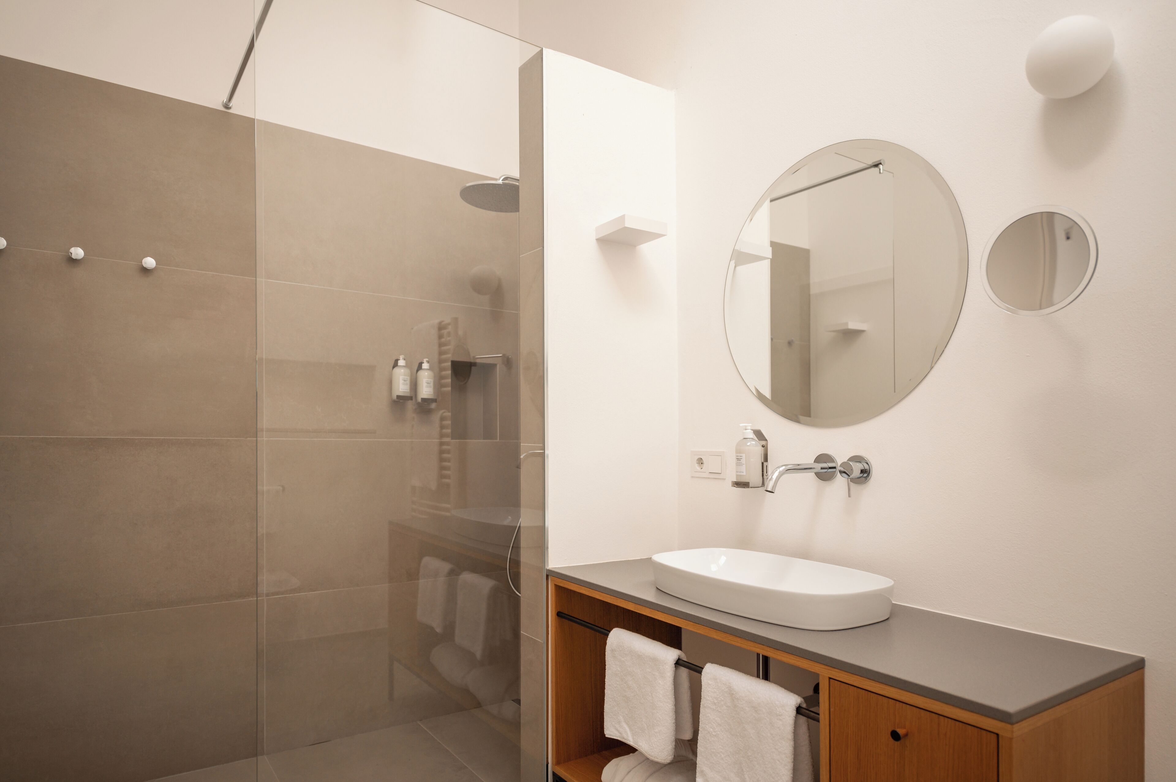 Une salle de bain moderne avec une douche à parois de verre, un miroir rond et un meuble-lavabo en bois avec un évier.