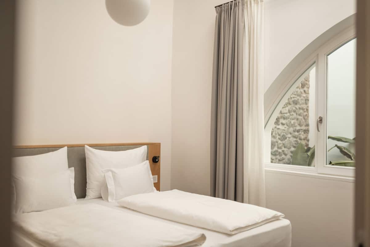 A neatly made bed with white linens in a hotel room with a stone wall and a window with curtains.