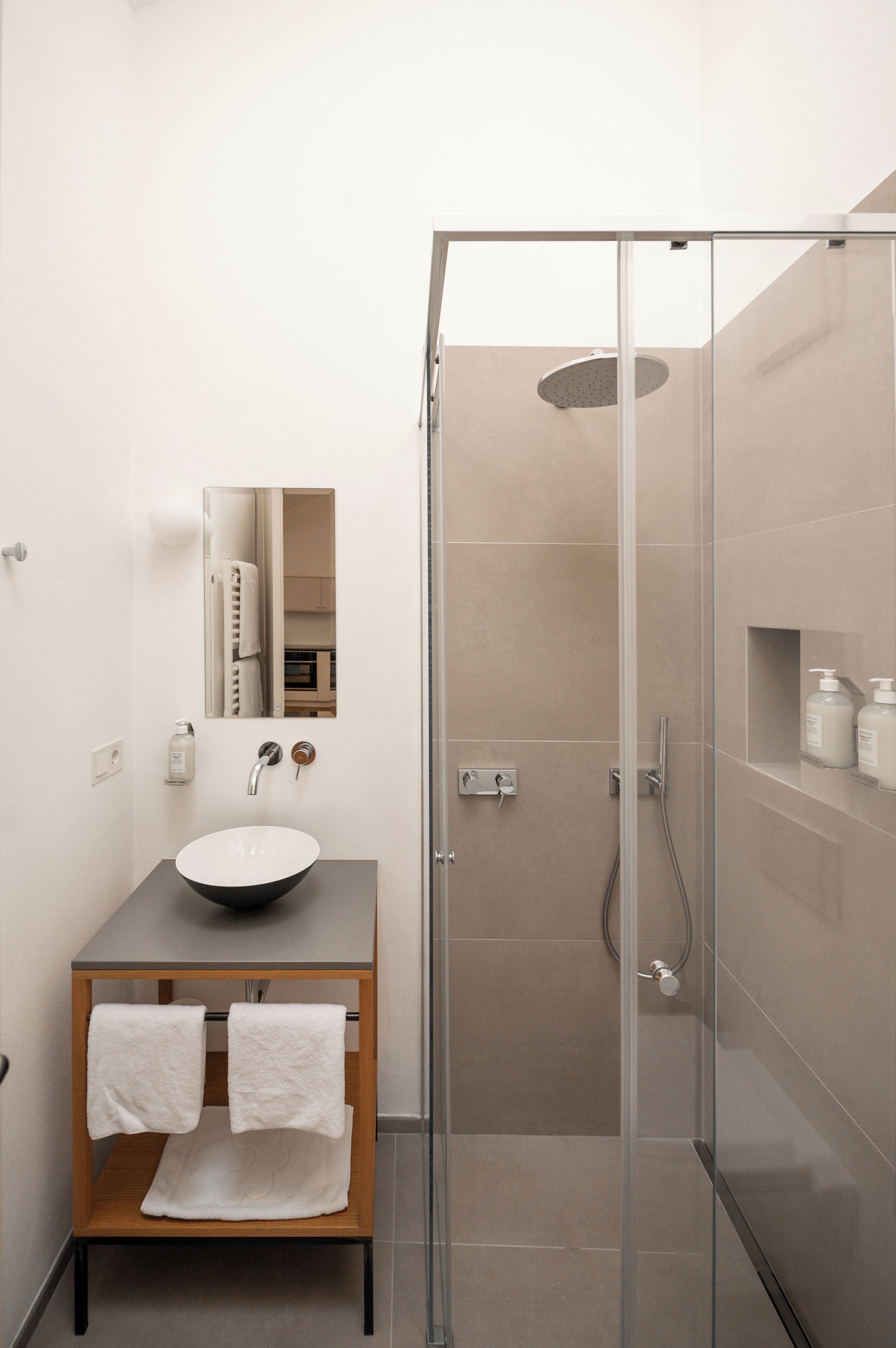 Un baño moderno con ducha envuelta en vidrio, tocador de madera con lavamanos blanco y espejo sobre el tocador.