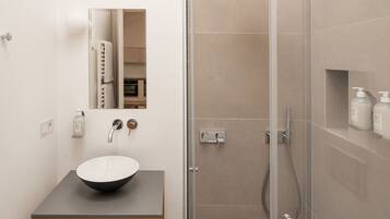 Un baño moderno con ducha envuelta en vidrio, tocador de madera con lavamanos blanco y espejo sobre el tocador.