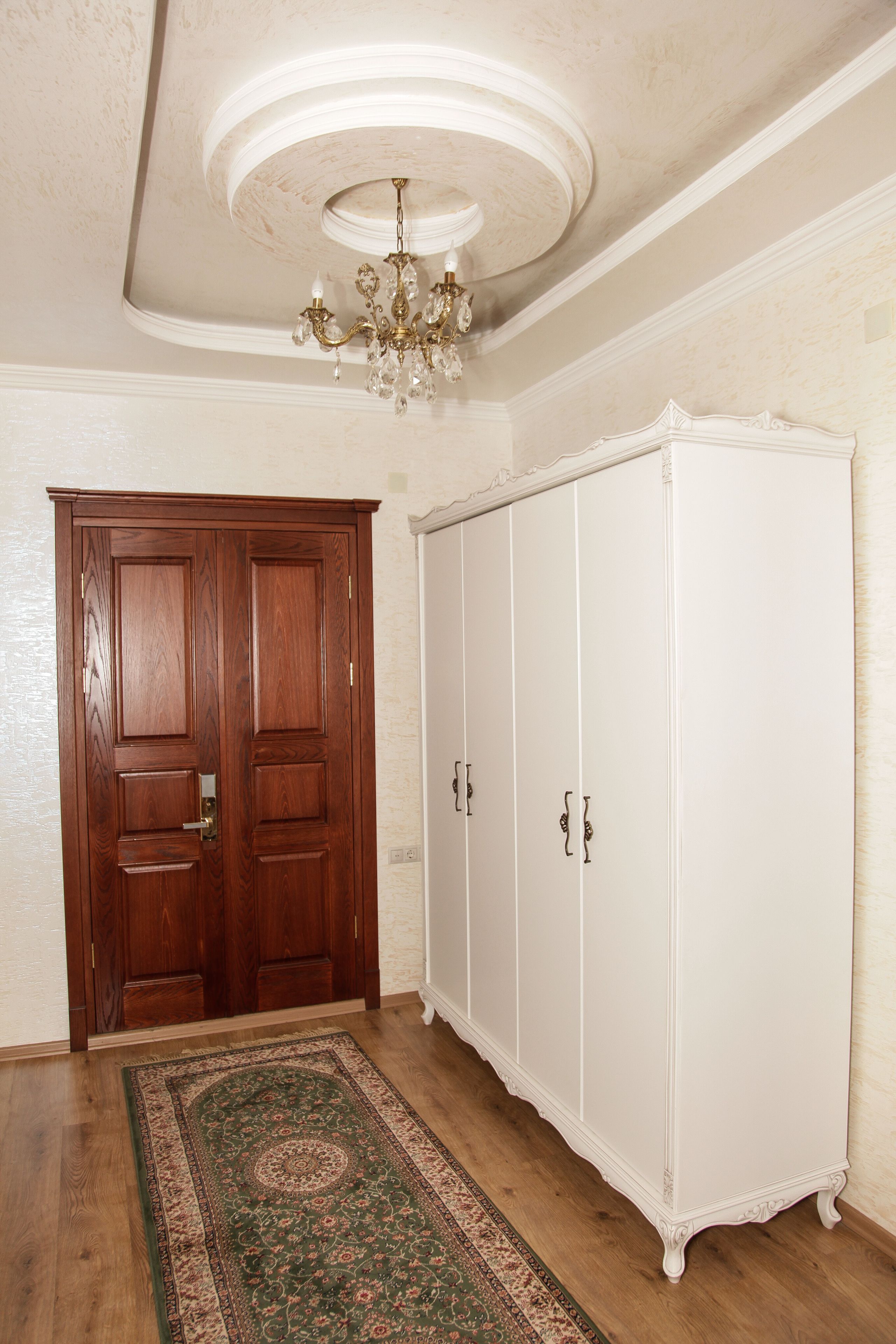 Una habitación con puerta de madera, armario blanco, alfombra estampada y una araña.