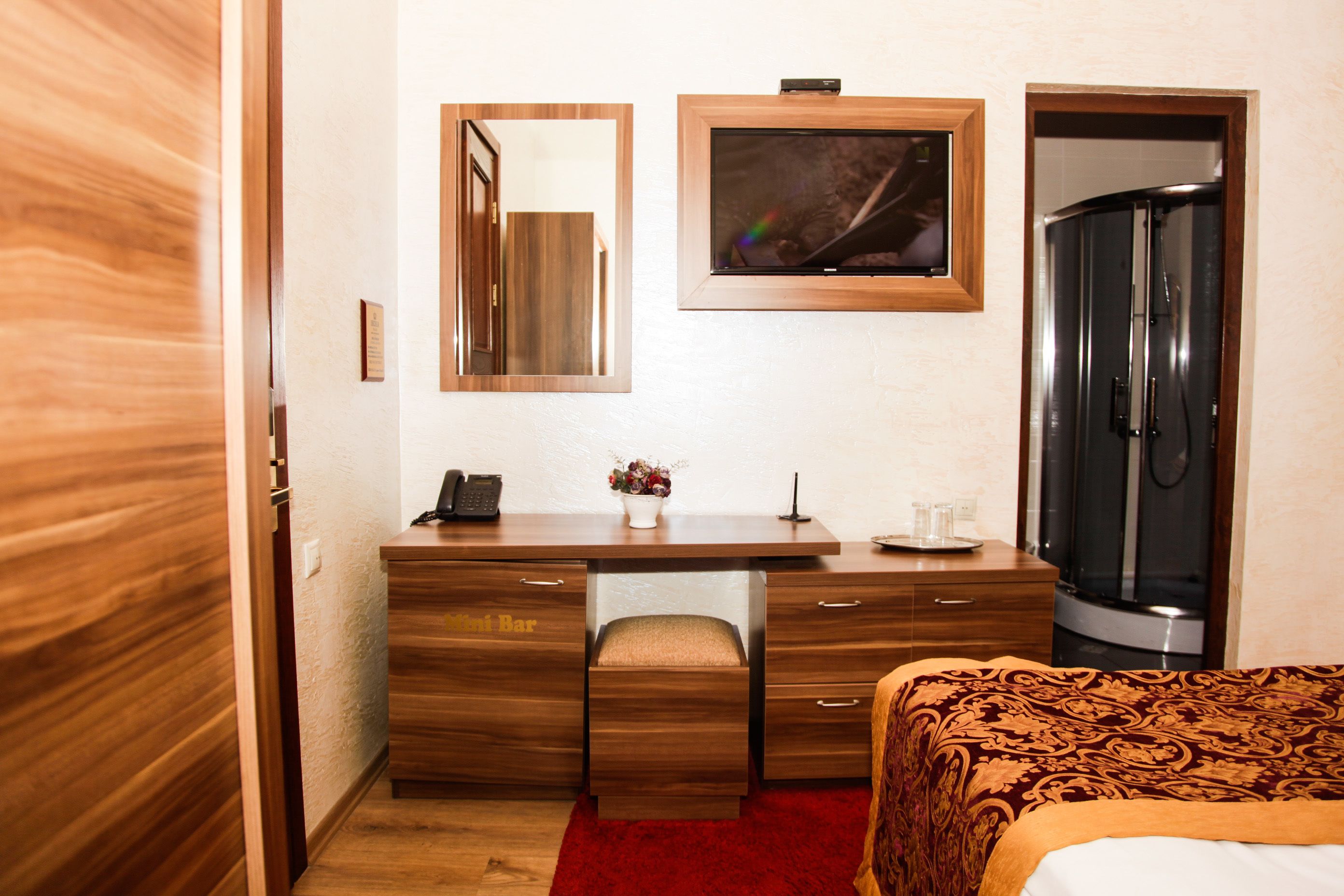 Een hotelkamer met een houten bureau, een bed met een beddeken met dessin, een spiegel, een lijst met afbeelding en een kledingkast.