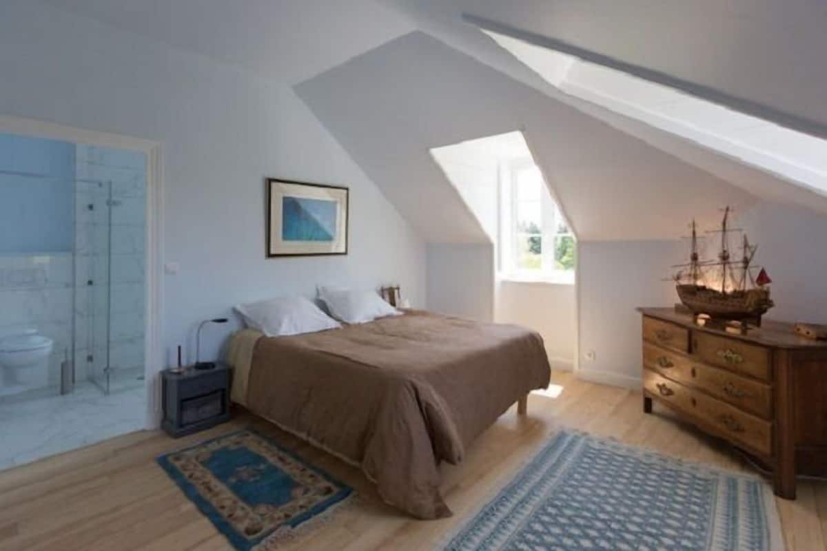 Une chambre avec un plafond en pente, un lit recouvert d’une couverture marron, une commode en bois, une maquette de bateau et un tableau encadré accroché au mur.