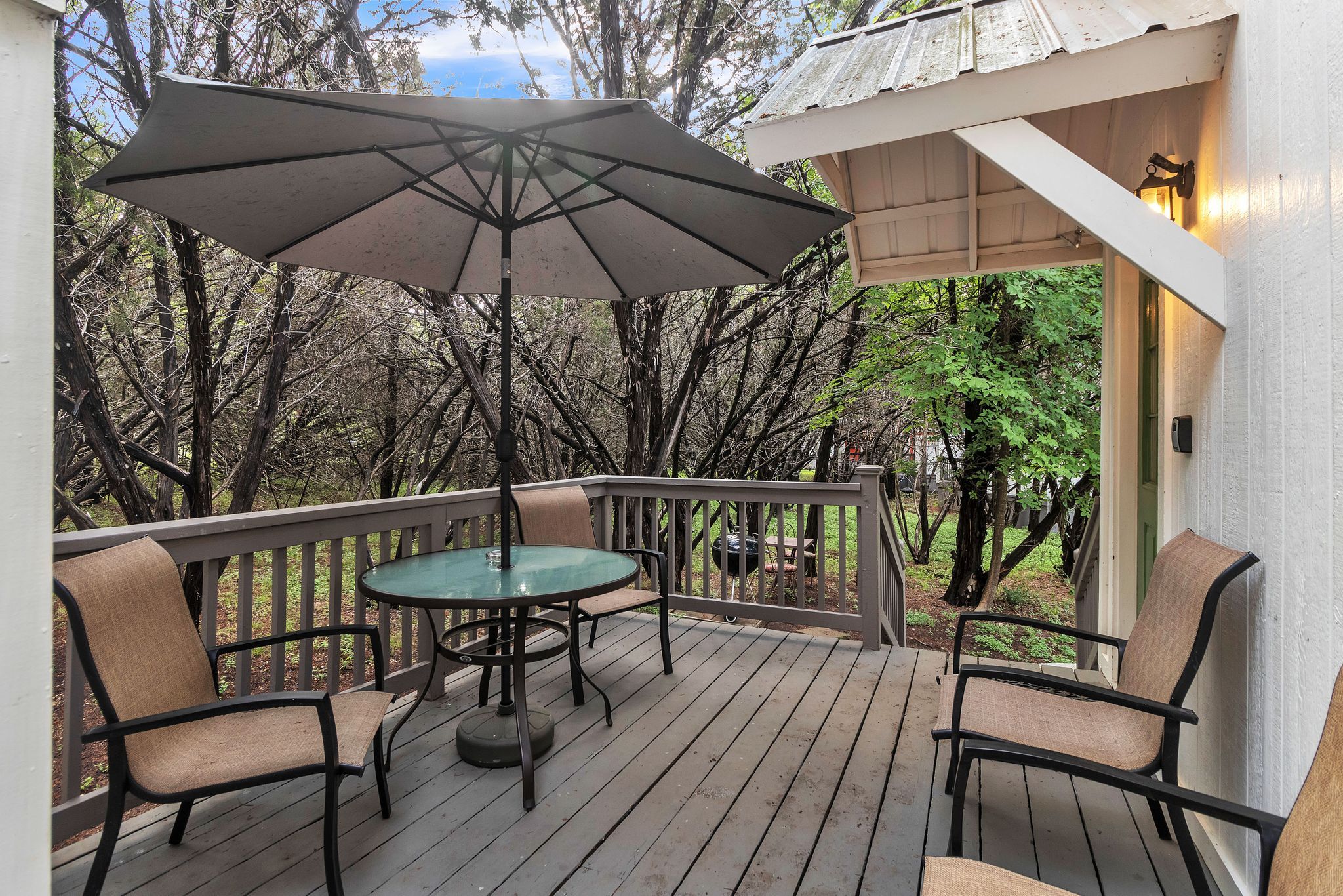 A wooden deck with a glass table and chairs, an umbrella, and a view of trees.