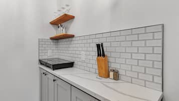 Una cocina con armarios blancos, encimera blanca, salpicadero con diseño de ladrillo, estante de madera con vasos, bloque de cuchillos y saleros.
