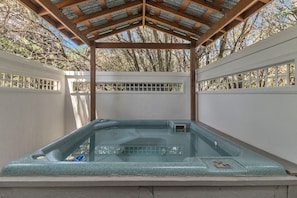 Un jacuzzi bajo una pérgola de madera con una valla blanca y árboles al fondo.