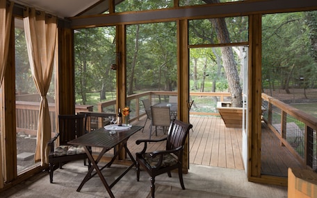 A covered outdoor deck with a wooden table and chairs, surrounded by large windows offering a view of a wooded area.