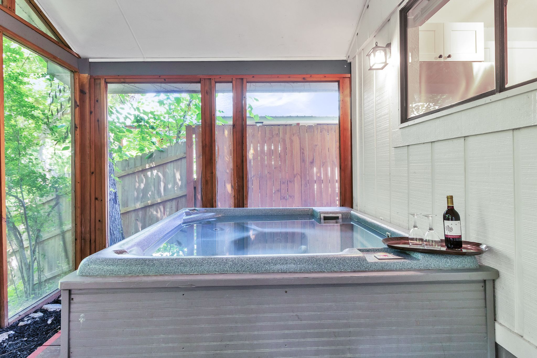 A hot tub with a wooden deck and a glass enclosure, a bottle of wine, and a glass on a tray.