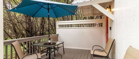 A wooden deck with a table and chairs, a blue umbrella, and a white wall with a window.