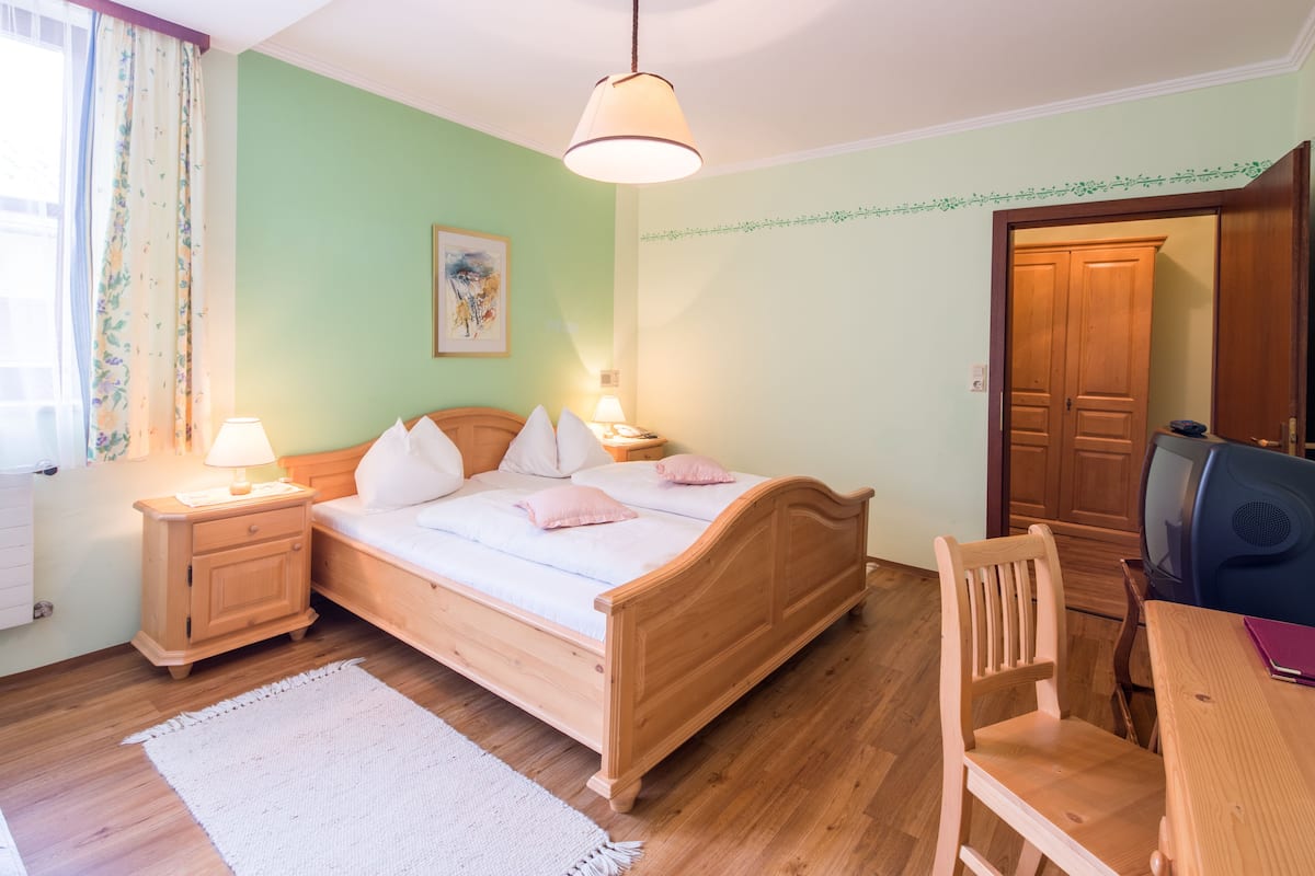 A bedroom with a wooden bed, bedside tables with lamps, a desk with a chair, and a television.
