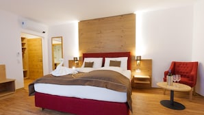 A modern hotel room with a large bed, wooden headboard, bedside tables with lamps, a red armchair, and a small round table with a bottle and glass. - Hotel Restaurant Kollar Göbl (Deutschlandsberg)