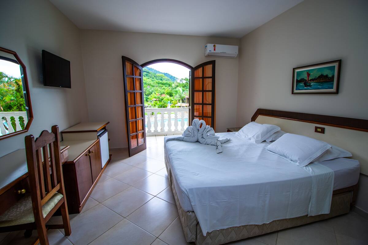 Une chambre d’hôtel équipée d’un lit, d’un bureau, d’une chaise et d’un balcon donnant sur un espace verdoyant.