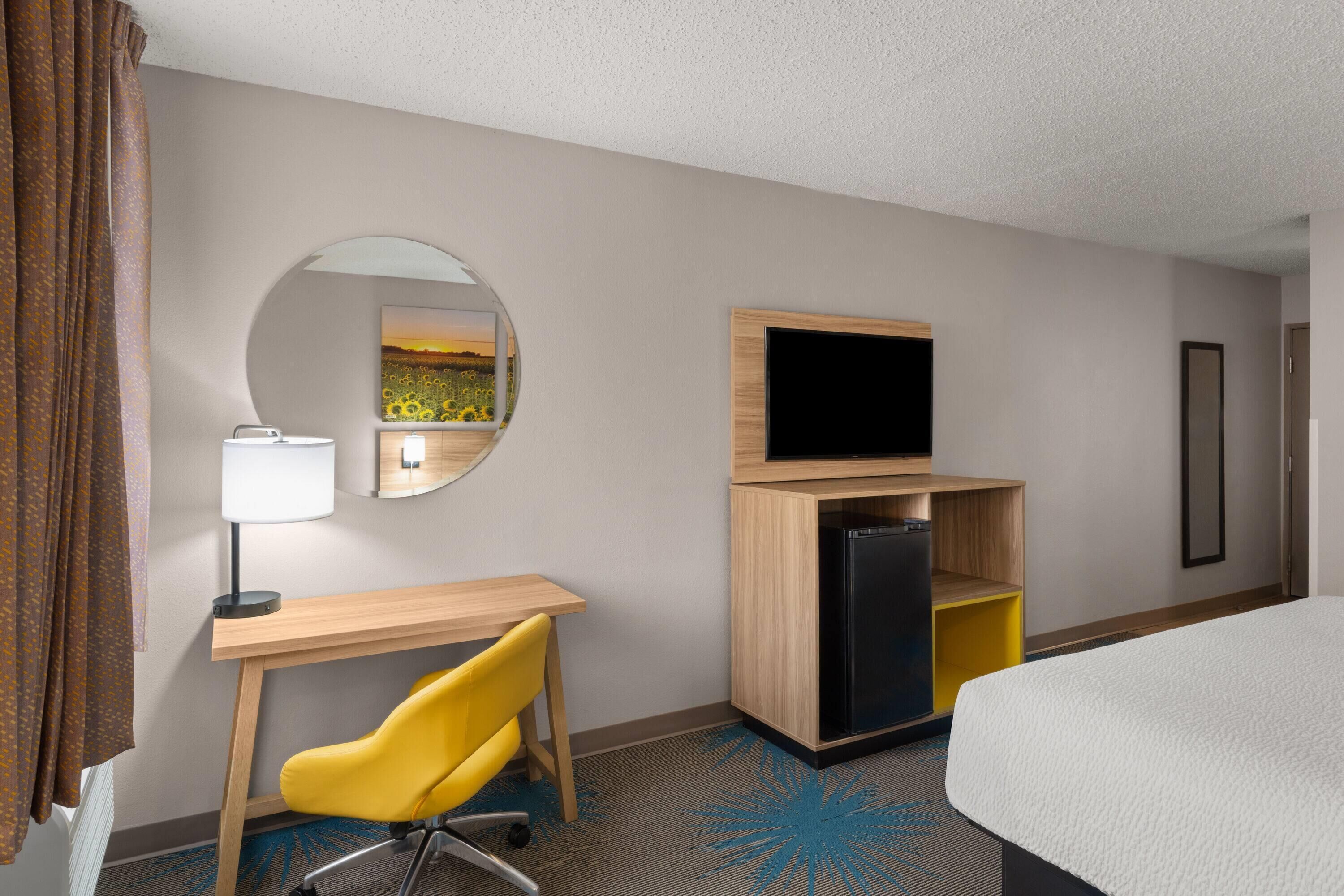 A hotel room with a bed, a desk with a chair, a TV, a mirror, and a refrigerator.