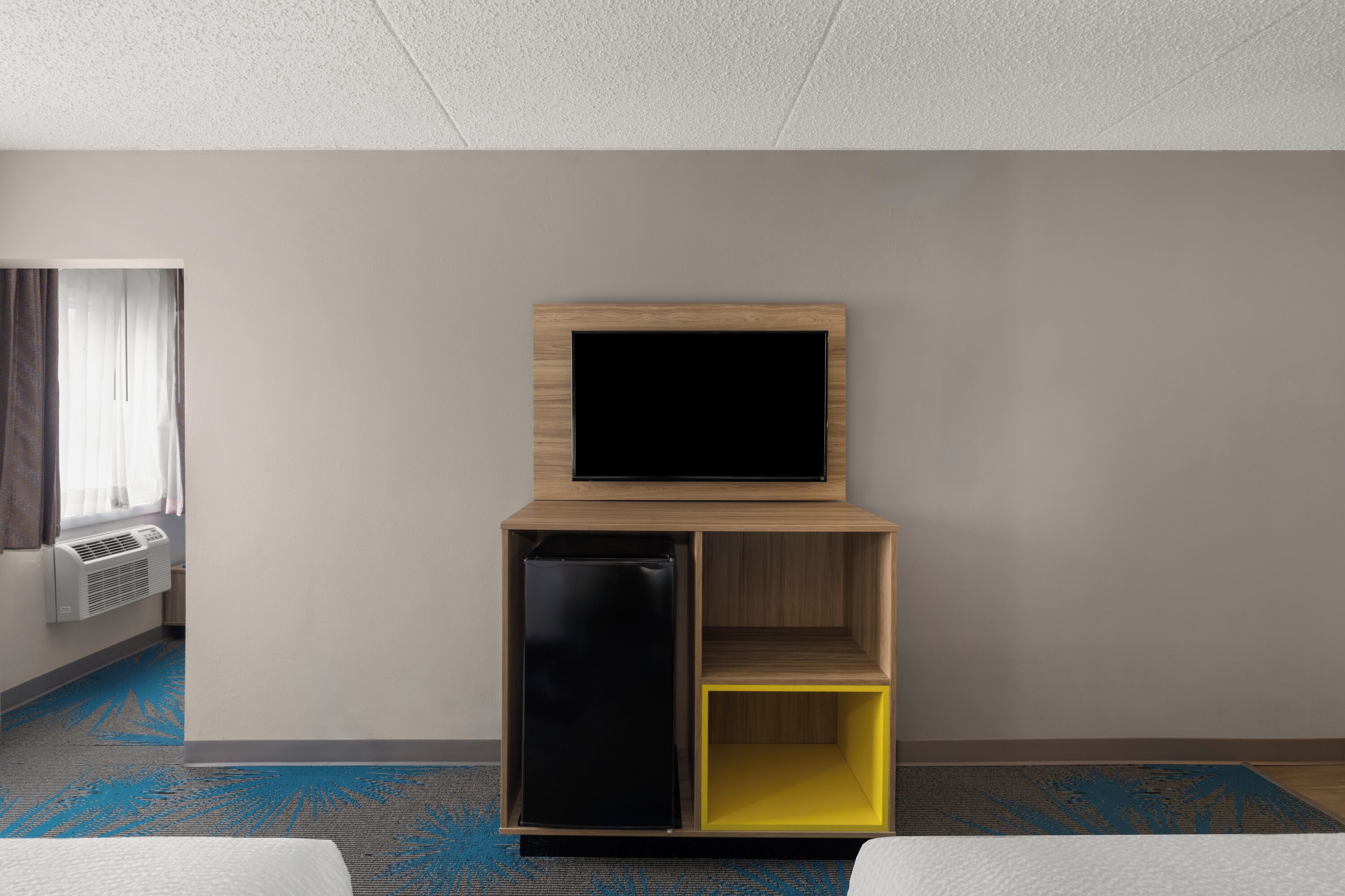 A hotel room with a flat-screen TV mounted on a wooden shelf, a mini-fridge, and a yellow storage box.