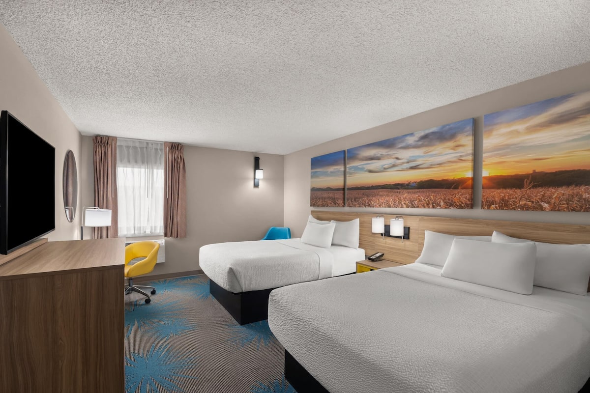 A hotel room with two beds, a desk, a TV, and a large window with a sunset view.