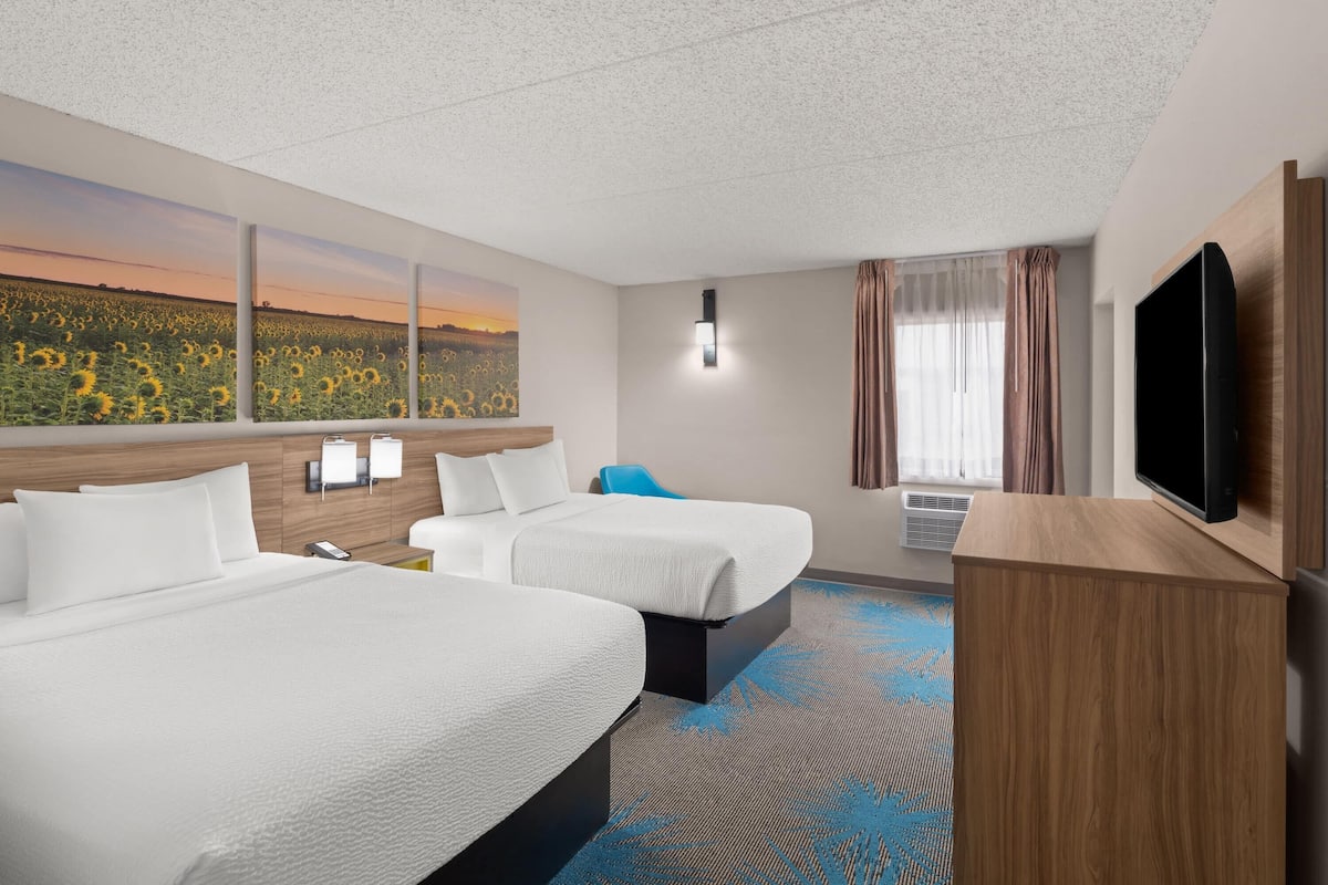 A hotel room with two beds, a television, a painting of a sunflower field, and a window with curtains.