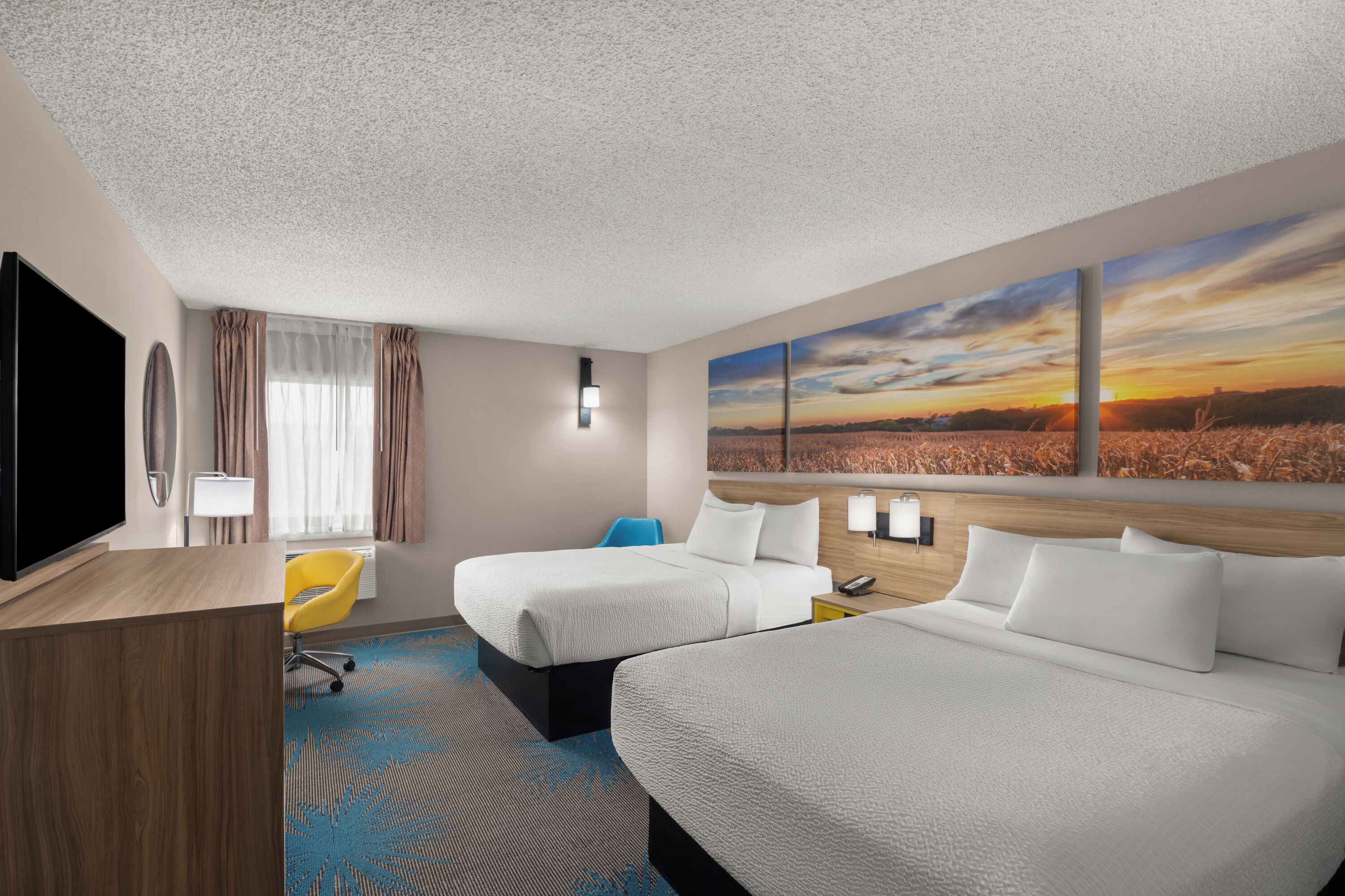 A hotel room with two beds, a desk, a TV, and a large window with a view of a sunset.