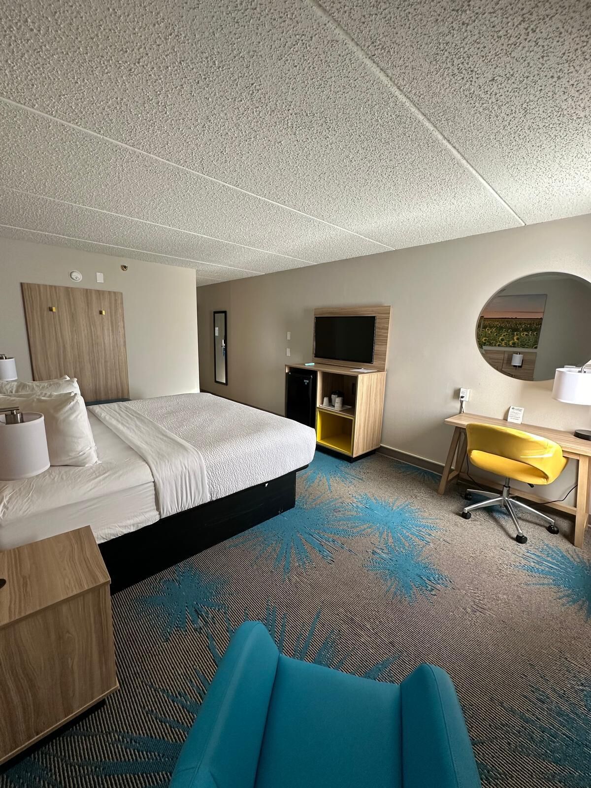 A hotel room with a bed, a desk, a chair, a TV, a mirror, and a small kitchenette.
