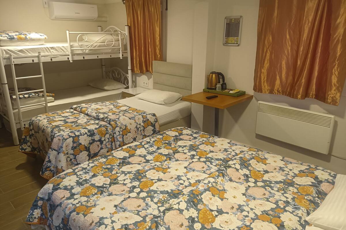 Une chambre de dortoir avec des lits superposés, deux lits simples avec une literie à motifs floraux, une petite table avec une bouilloire et un climatiseur fixé au mur.