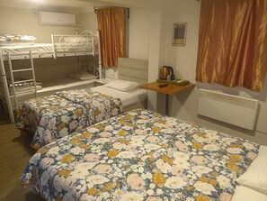 Een studentenkamer met stapelbedden, twee enkele bedden met bloemenbeddengoed, een tafeltje met een waterkoker en een wandairconditioner.