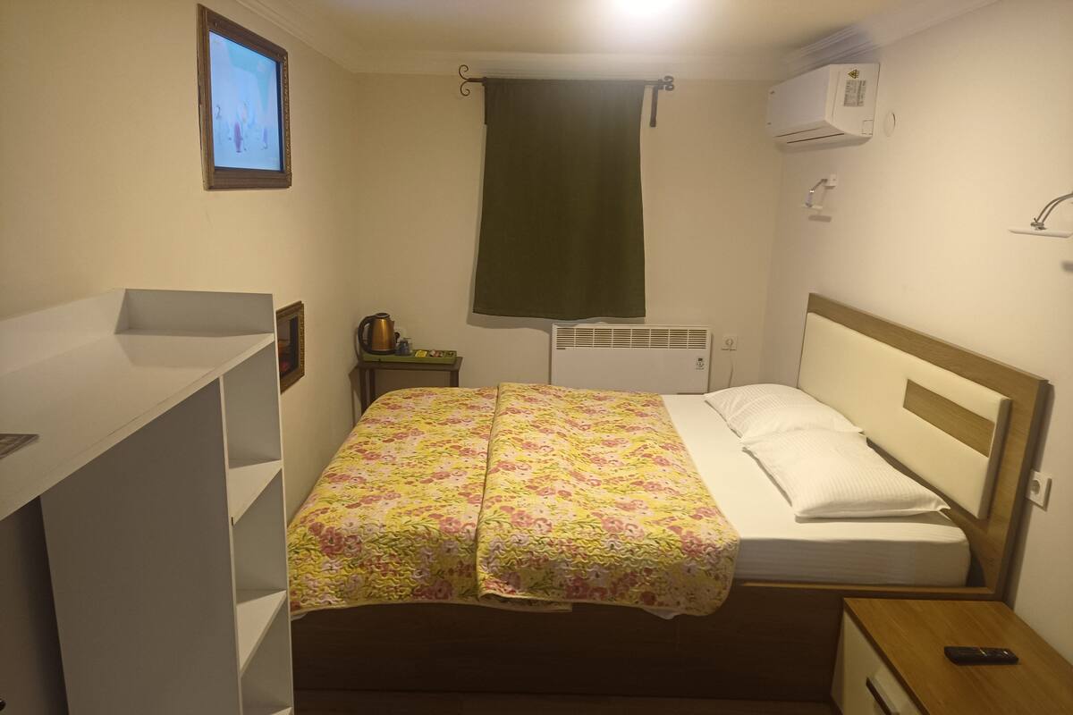 Une petite chambre à coucher, bien rangée, avec un lit, une table de chevet, une commode, un radiateur fixé au mur et un téléviseur accroché au mur.