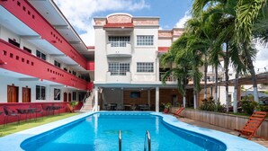 Un espace piscine avec une piscine bleue, entouré d’un bâtiment rouge et blanc doté de nombreux balcons et d’un espace salon extérieur.
