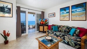 A living room with a sofa, a coffee table, and a TV. There are framed pictures on the wall and a view of the beach through the glass door.