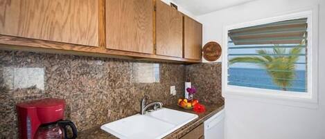 A kitchen with granite countertops, wooden cabinets, a red coffee maker, a white sink, and a window with a view of the ocean.