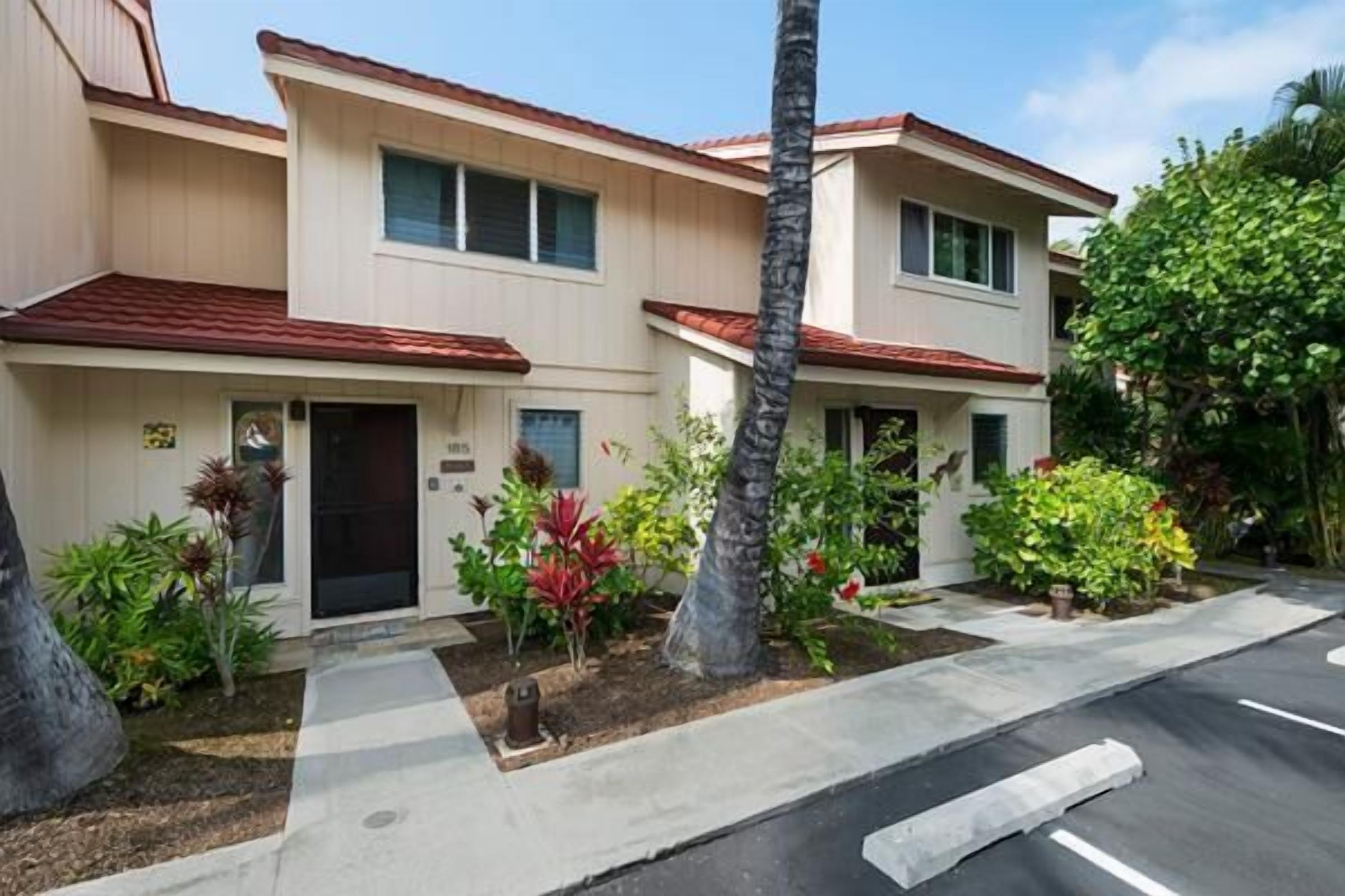 A two-story residential building with a red-tiled roof, a front entrance, and a small garden with various plants and a palm tree.