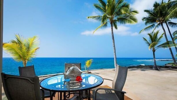A round glass table with chairs, a fish sculpture, and a red flower on a balcony overlooking the ocean with palm trees.