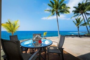 A round glass table with chairs, a fish sculpture, and a red flower on a balcony overlooking the ocean with palm trees. - Keauhou Kona Surf and Racquet Club Townhouse# 185 (Kailua-Kona)