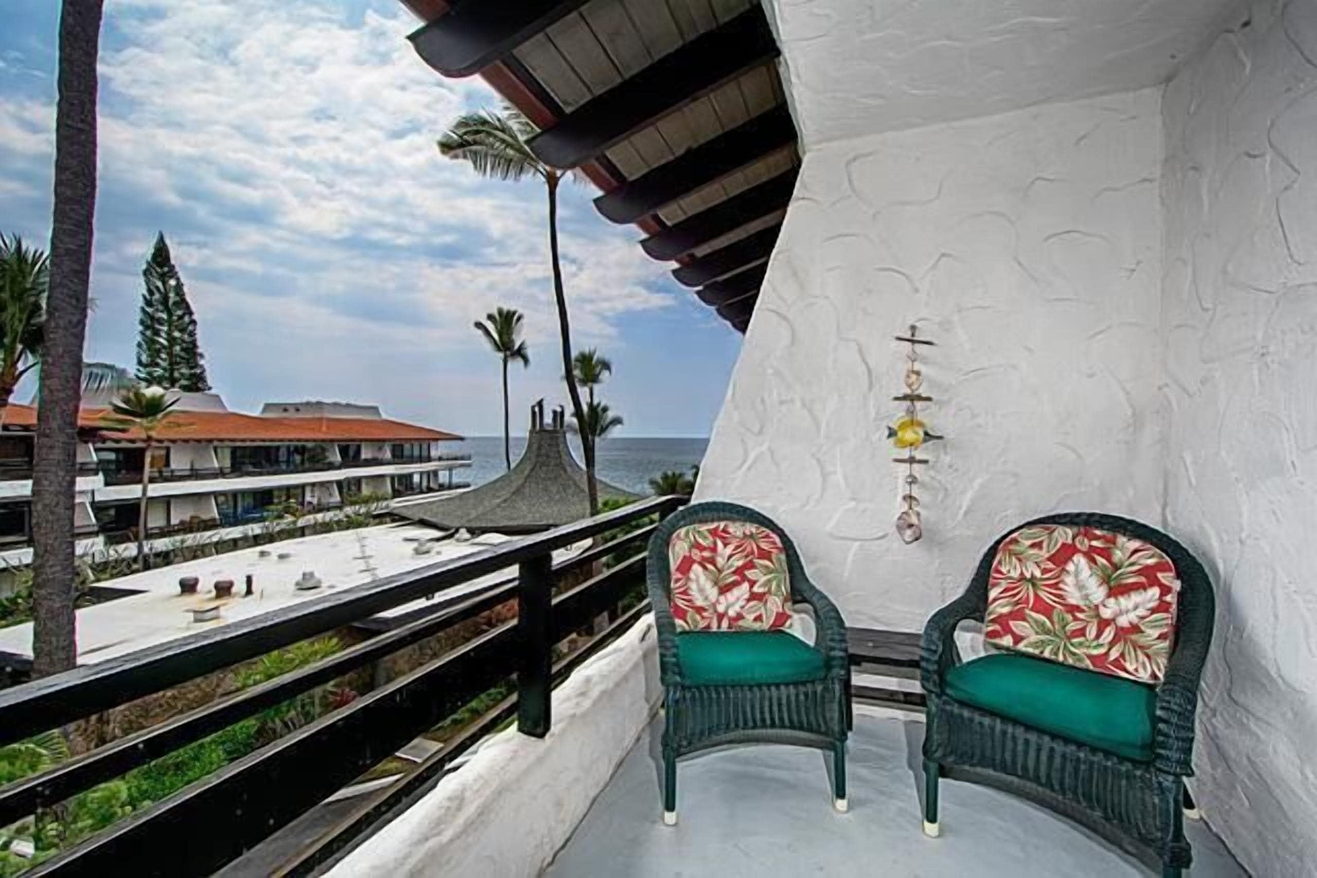A balcony with two wicker chairs, a white textured wall, and a view of the ocean and buildings.