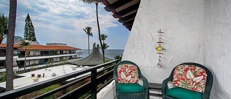 A balcony with two wicker chairs, a white textured wall, and a view of the ocean and buildings.