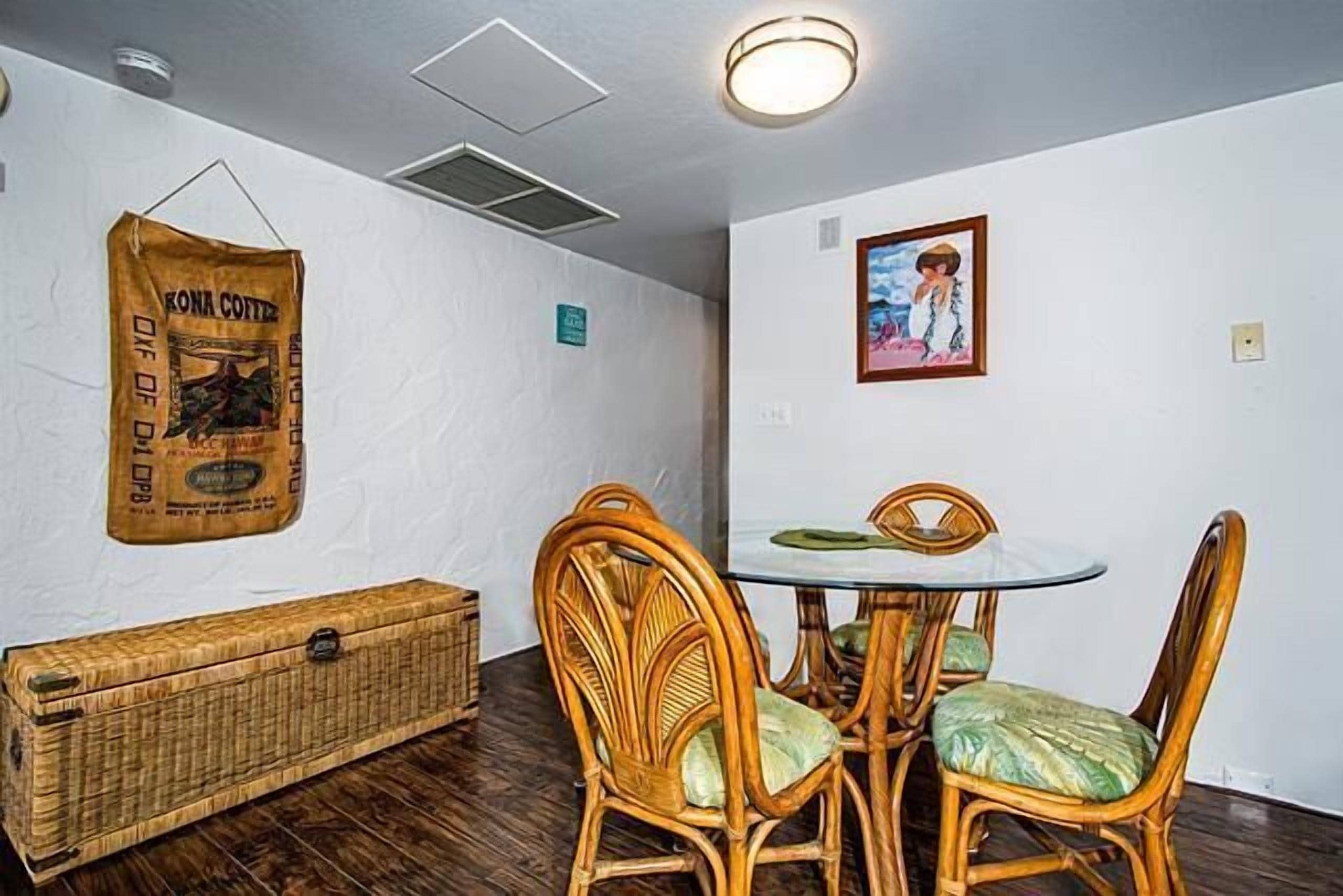 Un comedor con una mesa redonda y cuatro sillas de mimbre, un cofre de mimbre y un cartel en la pared.