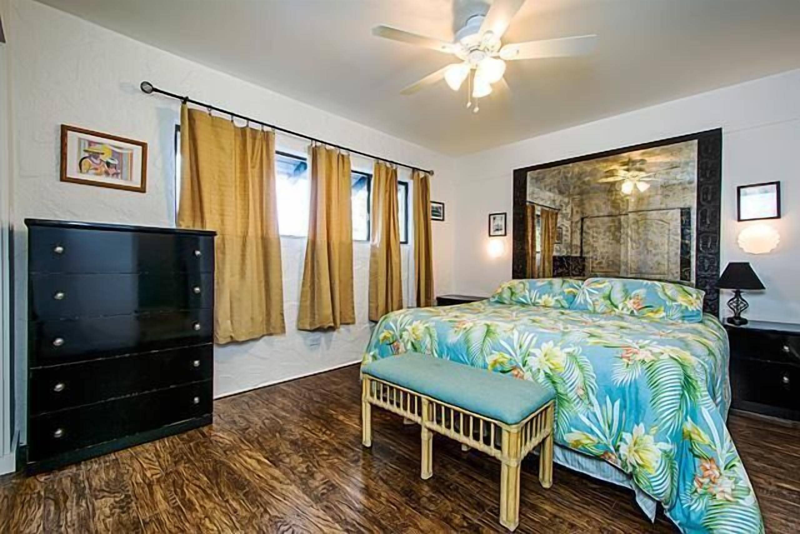 Ein Schlafzimmer mit einem Bett, einer hölzernen Bank, einem Schminktisch, einem Deckenventilator und einem großen Spiegel.