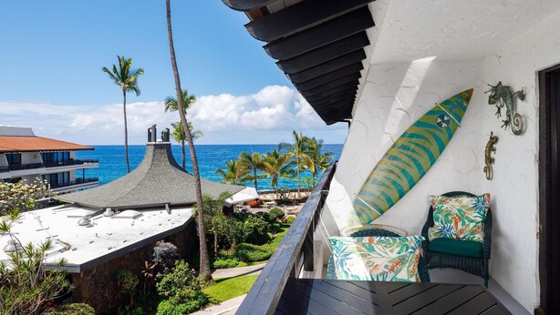 A balcony with a view of the ocean, palm trees, and a surfboard decoration.