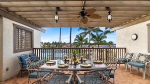 A covered outdoor dining area with a table set for four, surrounded by wicker chairs with blue cushions. A ceiling fan and two wall-mounted lights are visible above the table. - Kona Coast Resort at Keauhou Gardens 8204 (Kailua-Kona)