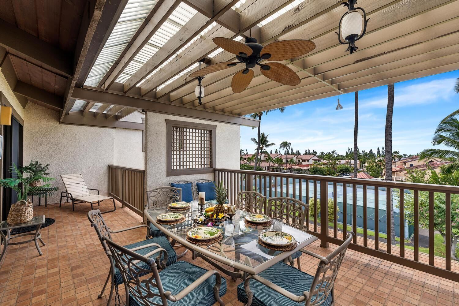 A covered outdoor dining area with a table set for two, surrounded by chairs with patterned cushions, a ceiling fan, and a view of a residential area with palm trees.