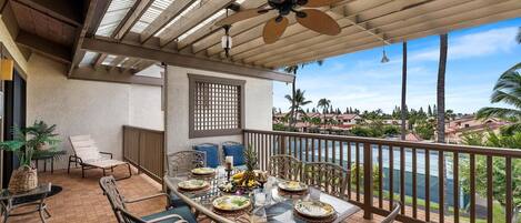 A covered outdoor dining area with a table set for two, surrounded by chairs with patterned cushions, a ceiling fan, and a view of a residential area with palm trees.
