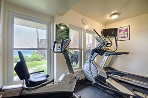 A gym with two treadmills and an elliptical machine, large windows with a view of greenery and a building, and a clock on the wall.