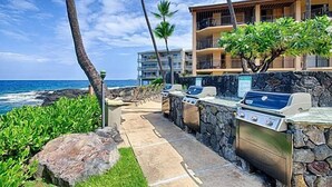 Ett utomhusområde med grillplats, omgivet av stenmurar och med grillar, med utsikt över havet och en byggnad med balkonger.