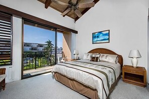 A bedroom with a large bed, a ceiling fan, a balcony with a view of buildings and palm trees, and a painting on the wall.