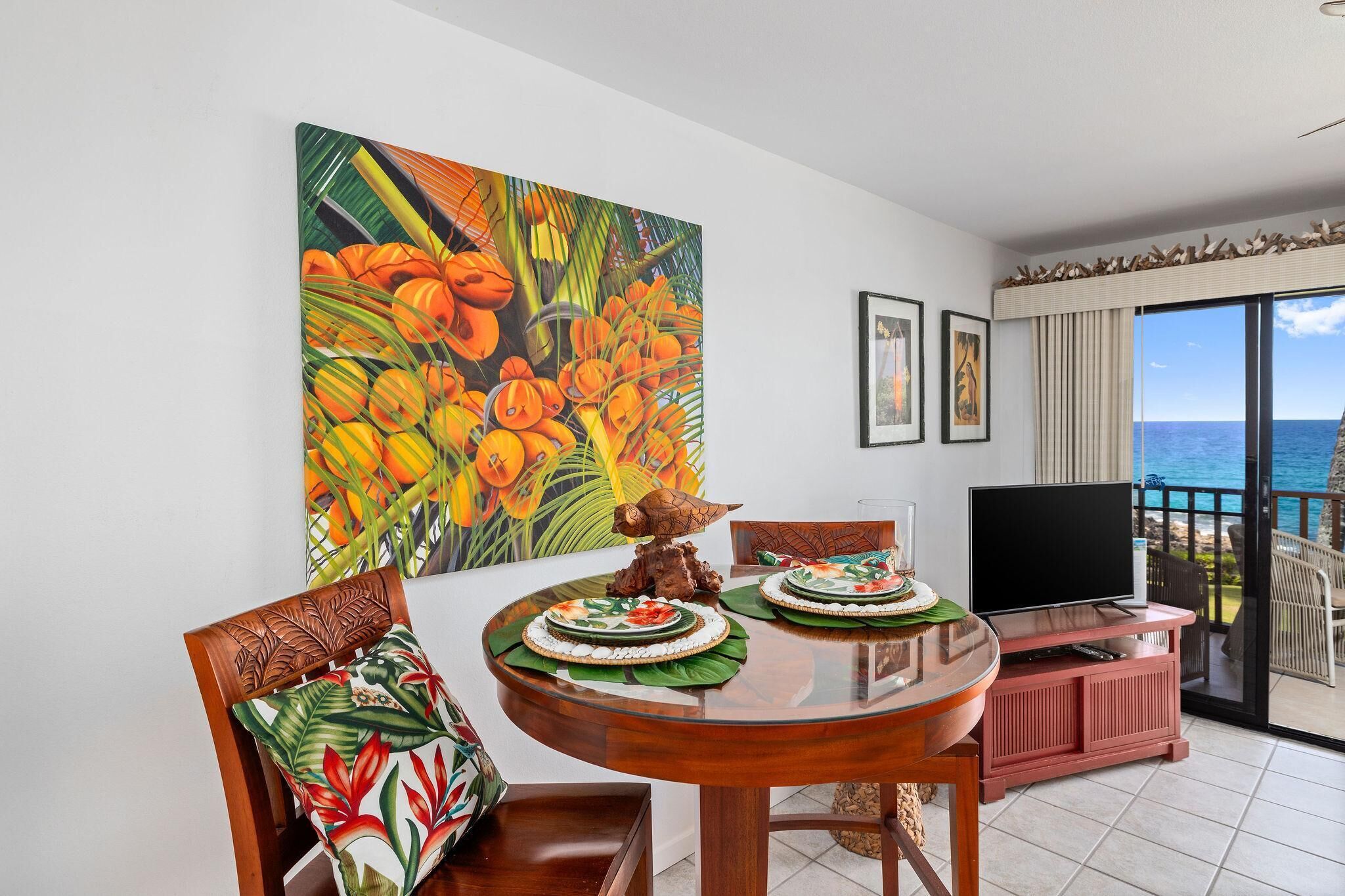 A dining area with a round table, chairs, and a TV. There is a large painting on the wall and a view of the ocean through the balcony.