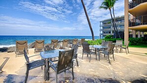 Un patio frente a la playa con mesas y sillas, con vista al océano, y un edificio y palmeras al fondo.