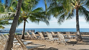A beach with rows of sun loungers, palm trees, and a view of the ocean. - Casa De Emdeko 204 (Kailua-Kona)
