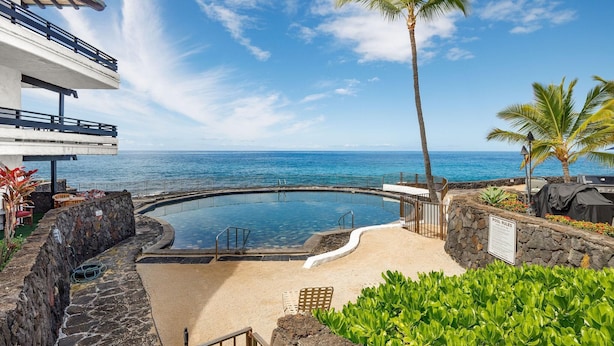 A pool area with a view of the ocean, surrounded by palm trees and a stone wall. - Casa De Emdeko 204 (Kailua-Kona)